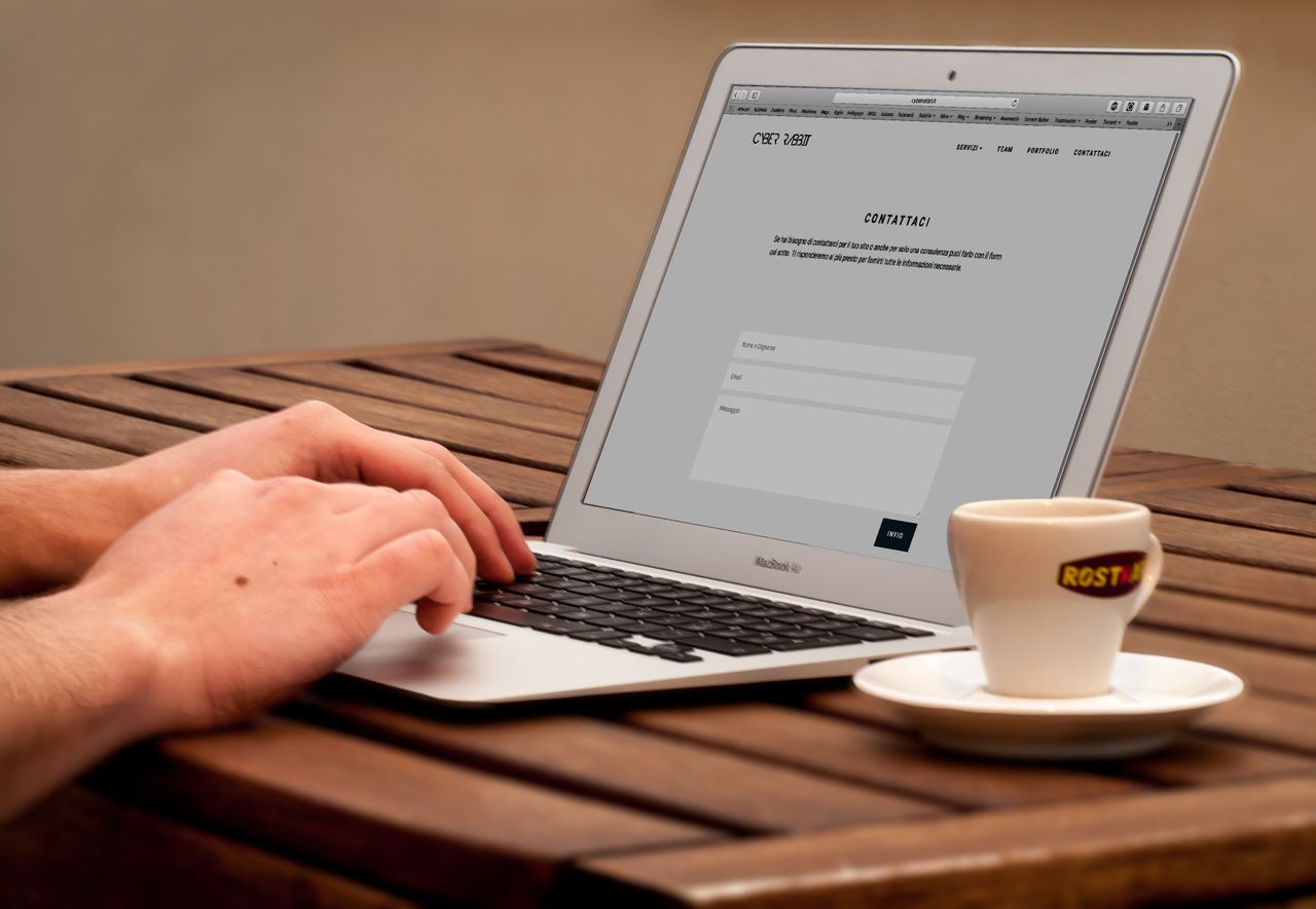 Gros plan sur des mains en train de taper sur un ordinateur portable, à côté d’une tasse de café posée sur un bureau en bois.
